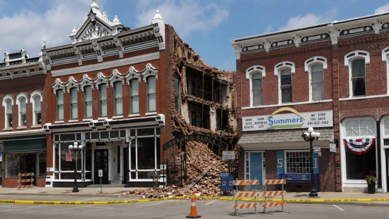 A historic red-brick building in downtown Albia with one side partially collapsed, exposing interior floors and wooden framing behind caution tape and barricades.