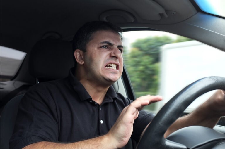 An angry driver grips the steering wheel and grimaces while looking out the windshield of a moving car.